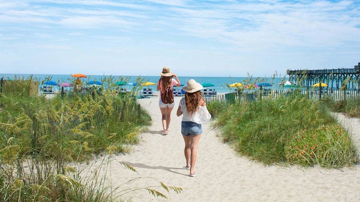 women with hats walking along myrtle beach with pier in background
